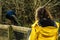 Colorful male peacock on a wooden fence. Young slim teenager girl in yellow jacket looking at the bird, back to viewer. Scene in a