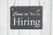 Come in we`re hiring text on a hanging chalkboard on weathered whitewash textured wood