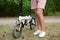 Compact folding bike next to the legs of an adult woman. A woman is standing in beige shorts and white sneakers