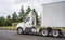 Compact powerful big rig white day cab semi truck with dry van semi trailer running on the wide highway road with trees on the
