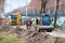 A compact road grader moves along a sidewalk under construction in front of a construction site and a team of road workers in blur