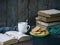 The composition of a stack of old books, open book, tea cups, glasses and plates of sugar cookies on a wooden background.