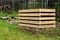 A composter made of natural beech prisms built horizontally with black foil inside in a meadow near a dark wooden fence.