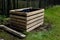 A composter made of natural beech prisms built horizontally with black foil inside in a meadow near a dark wooden fence.