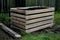 A composter made of natural beech prisms built horizontally with black foil inside in a meadow near a dark wooden fence.