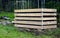 A composter made of natural beech prisms built horizontally with black foil inside in a meadow near a dark wooden fence.