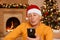 Concentrated attentive mature man wearing orange sweater and santa claus hat using mobile phone while posing at home in decorated