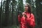 Concentrated girl in a red jacket stands in the evening in the woods with a smartphone in her hands and looks away anxiously. Girl