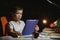 Concentrated schoolboy reading book at table with books, plant, lamp, colour pencils, apple, and textbook