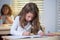 Concentrated schoolgirl sitting at desk and writing in exercise book with classmate sitting behind.