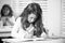 Concentrated schoolgirl sitting at desk and writing in exercise book with classmate sitting behind.