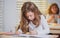 Concentrated schoolgirl sitting at desk and writing in exercise book with classmate sitting behind.