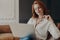 Concentrated serious redhead woman makes remote job indoors, sits in comfortable armchair, holds transparent glasses, reads