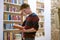 Concentrated student with glasses reading a book in library next to bookshelf. Stone wall in background