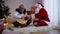 Concentrated woman with guitar sitting at New Year tree with man in Santa Clause costume teaching playing. Caucasian