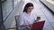 Concentrated woman in headphones surfing Internet on laptop sitting at table with refreshing fruit cocktail outdoors