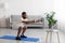 Concentrated young black bearded athlete man in white sports clothes squatting in living room interior, profile