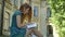 Concentrated young ginger female student reading book and sitting in park near college building, sunny day outside