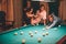 Concentrated young redhead man aiming into billiard ball. He look straight forward. Young woman in pink dress sit at billiard