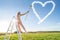 Conceptual photo of smiling woman on a ladder with brush. Happy girl painting white heart mark on a blue sky. Summer vibes. Love