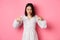 Concerned asian woman peeking down and pointing hand at disturbing banner, frowning upset, standing over pink background