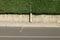 Concrete enclosure wall with green hedge above. A tiled sidewalk, cement pole and asphalt road in front.