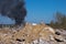 The concrete remains of a ruined building with protruding stilts against a background of black smoke against a blue sky