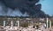 The concrete remains of a ruined building with protruding stilts against a background of black smoke against a blue sky