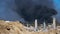 The concrete remains of a ruined building with protruding stilts against a background of black smoke against a blue sky