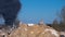 The concrete remains of a ruined building with protruding stilts against a background of black smoke against a blue sky