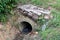 Concrete sewer pipe exit covered with red bricks and broken concrete used as small bridge surrounded with uncut grass and small