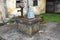 Concrete well with rusted metal cover and grey bucket locked with padlock next to old broken water pump in front of abandoned old