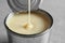 Condensed milk pouring into tin can on grey, closeup. Dairy product