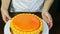 Confectioner rotates white spinning stand with orange glazed round cheesecake