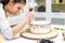 A confectioner squeezes liquid chocolate from a pastry bag onto a white cream biscuit cake on a wooden stand. The