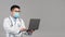 Confident adult chinese man doctor in white coat, glasses, protective mask typing on laptop