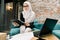 Confident Arabic female entrepreneur, wearing hijab and eyeglasses, sitting in the modern loft office room on sofa