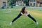 Confident athletic woman demonstrates her flexibility, stretches outdoors, does exercises early in morning on green grass near