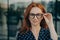 Confident beautiful red-haired business woman adjusting spectacles eyeglasses posing on street
