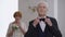 Confident Caucasian grey-haired groom adjusting bow tie looking at camera smiling. Blurred happy bride with bouquet at