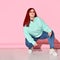 Confident chubby woman in jeans, bright sweater and stylish glasses poses squatting on floor.