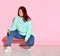 Confident chubby woman in jeans, bright sweater and stylish glasses poses squatting on floor. Full length portrait isolated on