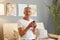Confident grey-haired mature man wearing casual white T-shirt sitting on sofa at home interior using mobile phone browsing