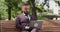 Confident mature businessman wearing suit working on laptop remotely, sitting on bench in park and drinking coffee