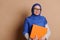 Confident mature Muslim female teacher in a hijab, looking at camera, posing with a stack of books, on beige background