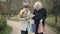 Confident senior Caucasian wife walking with husband in autumn park with bouquet of flowers and shopping bags and