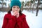 Confident serene brunette woman in bright red coat blowing on her crossed hands near her face while walking on the snowy forest