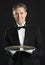 Confident Waiter In Tuxedo With Tray Standing Against Black Back