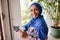 Confident young Arab Muslim pretty woman with covered head in blue hijab, smiles looking at camera, washing windows, removing