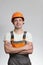 Confident young construction worker in hard hat and overalls with arms folded on the chest on grey studio background